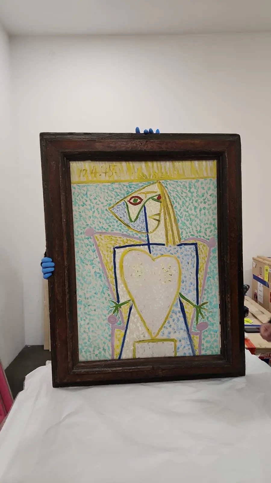 A framed painting held by a gloved hand against a white wall